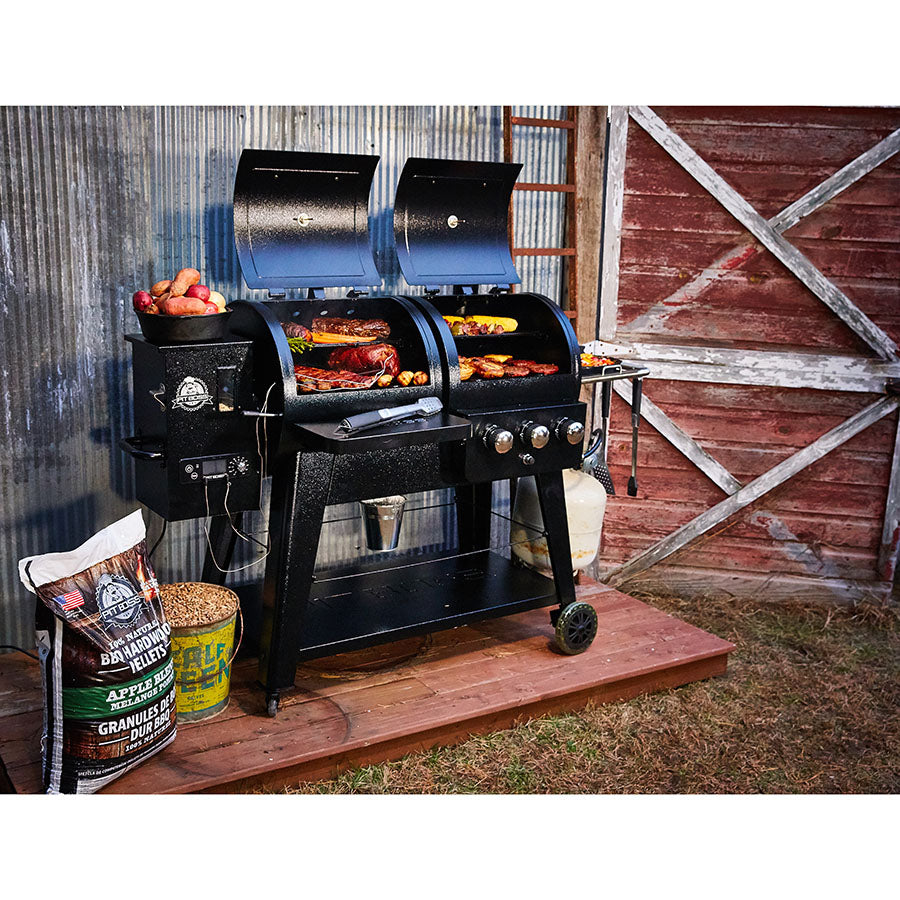 lifestyle_3, Black grill with silver accents and Pit Boss logo. Shown next to barn while meats and veggies are cooking