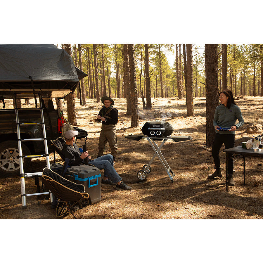 Grill is the forest with campers surrounding it and making a meal on it.