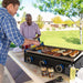 Men cooking on the griddle on a picni table with burgers and hot dogs being cooked. Pit Boss spices sitting next to the griddle.