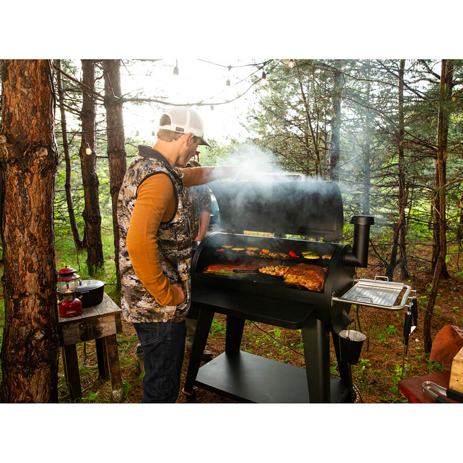 Man opening the hood to a Pit Boss Sportsman 1100 Grill with veggies and meats being smoked. Wilderness background