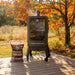 lifestyle_3, Black smoker with small Pit boss logo on transparent door and engraved "Pro Series" on front. Smoker on porch in front of autumn trees