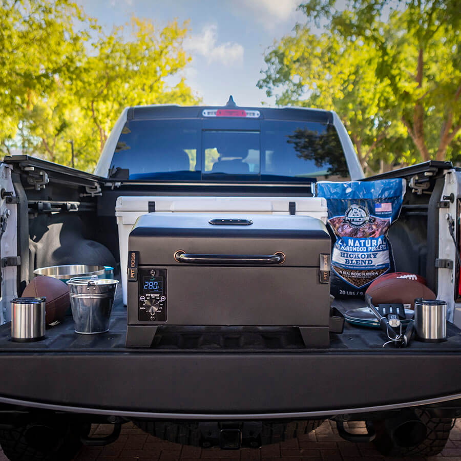 Grill in the back of a truck next to footballs, Pit Boss hardwood pellets and a cooler.