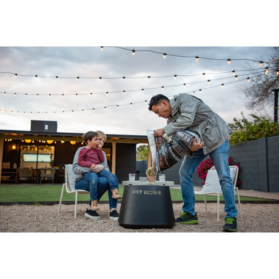 lifestlye_4, All black fire pit with silver lettering engraved "Pit Boss" logo. Shot of man pouring wood pellets into pit