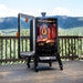 Smoker sitting on a deck with the mountains and trees in the background. Smoker door open with smoke poring out. Meats cooking on the racks and hanging