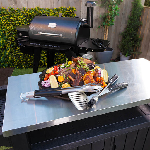 Grilling tools displayed next to veggies and meats on plate on table next to grill.