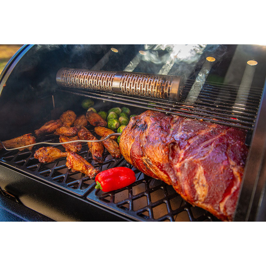 lifestyle_2, Silver, cylindrical smoker tube with a-maze-n logo engraved on end and holes for air. Pictured with pellets inside and inside grill with meat and veggies