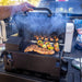 Man opening the hood of the grill. Meats and veggies cooking on both grilling racks. Smoke coming out of the grill. Grill in the back of a pick-up truck.