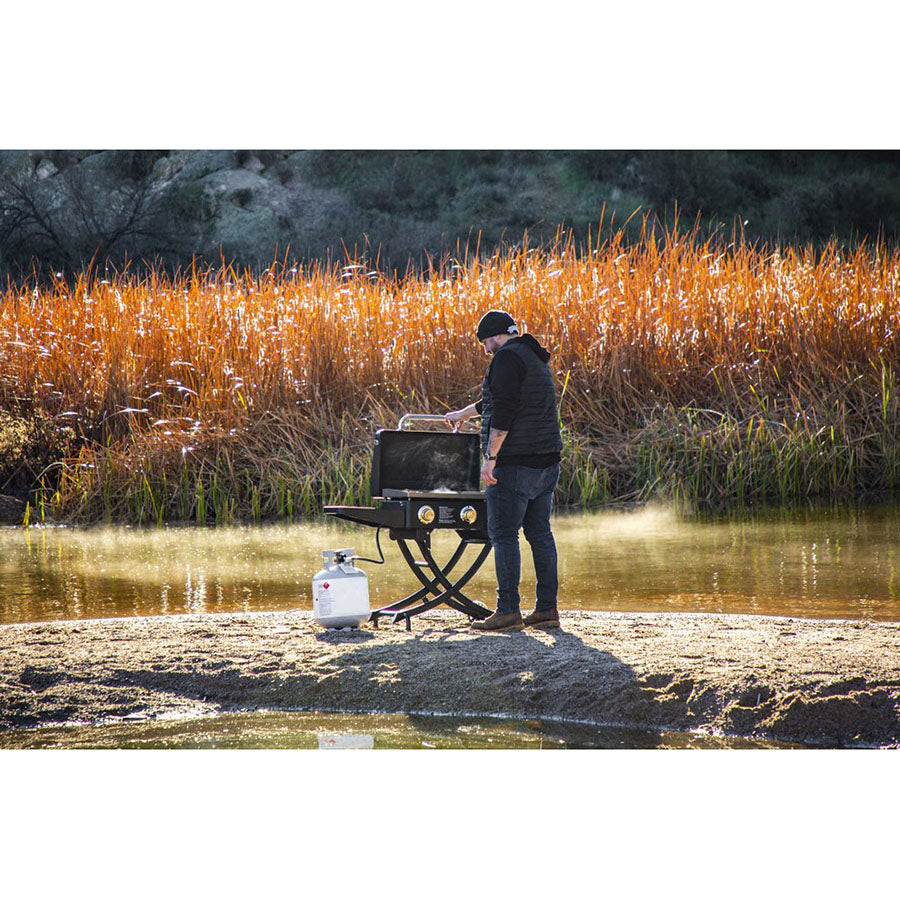 man opening griddle on river bank with reeds in background