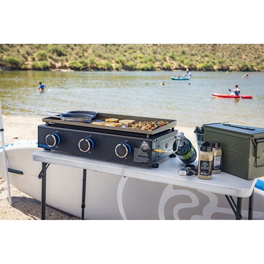 Griddle sitting on a foldable table next to Pit Boss spices and container around a lake.