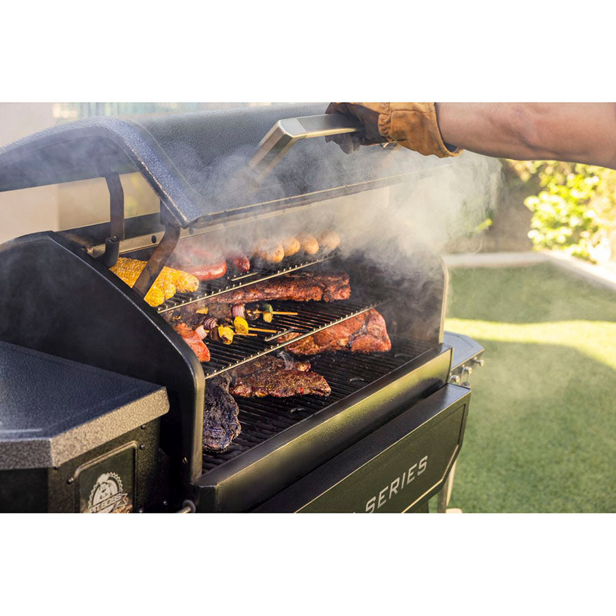 Close view of man opening the hood of the grill. Meats and veggies being cooked on all three racks of the grill. Smoke steaming out.