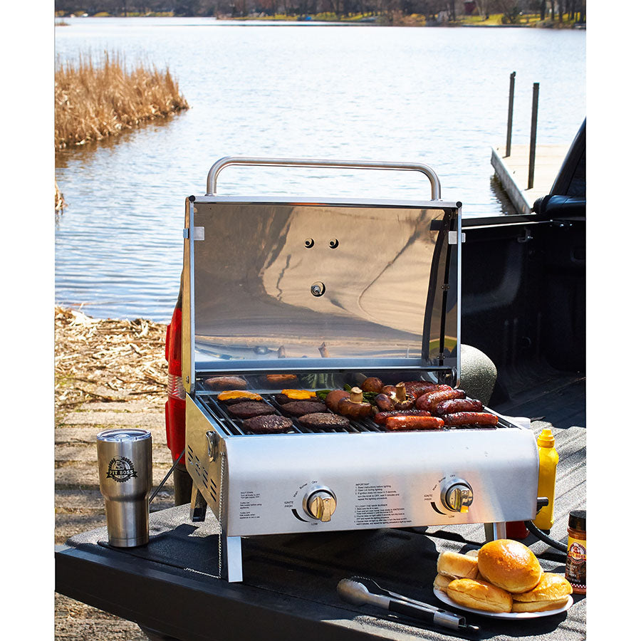 lifestyle_3, Silver/grey grill with black letter and a small Pit Boss logo. Grill on back of truck grilling meat an veggies near body of water