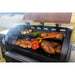 Close up of meats and veggies cooking on both grilling racks with smoke coming out. Grill outside in a yard.