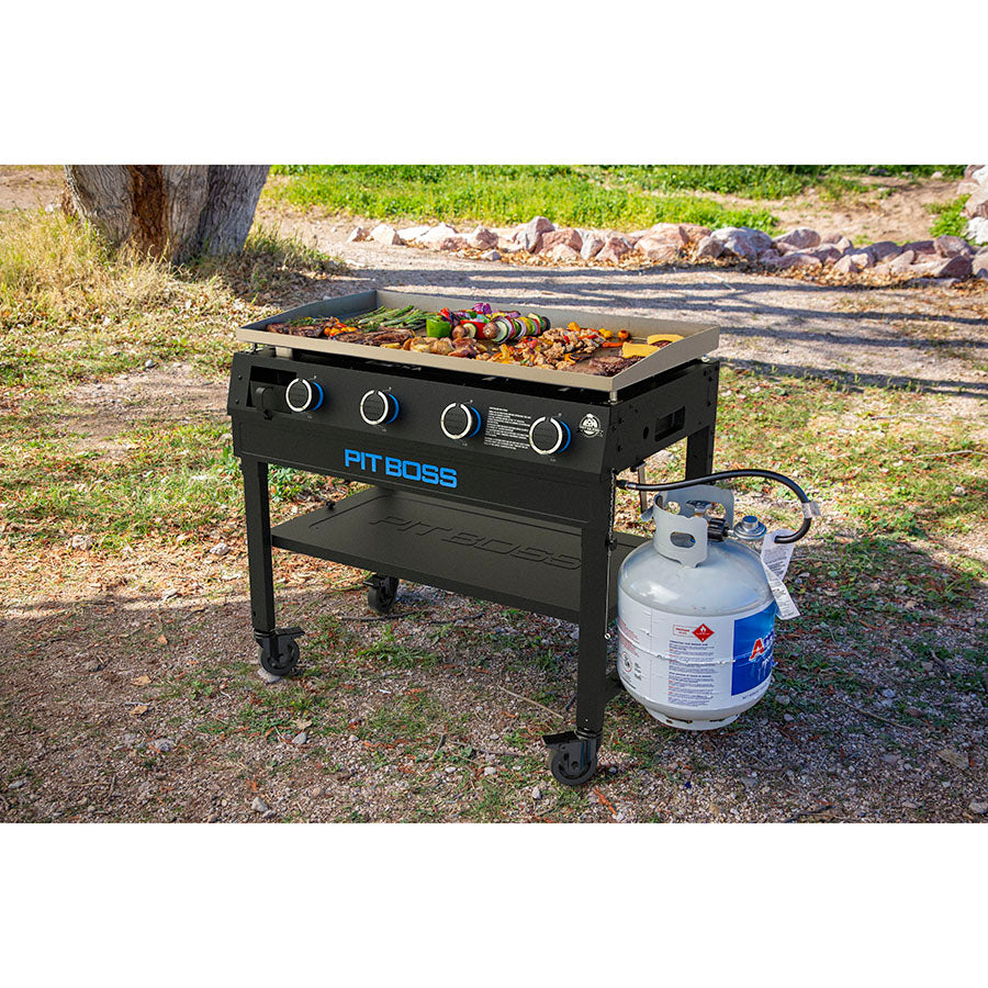 Griddle sitting in the ass with meats and veggies being cooked on it and propane tank plugged in.